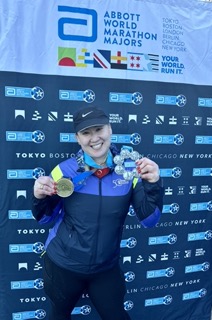 A smiling runner holds a gold medal and a medal on a ribbon, standing in front of a backdrop featuring marathon logos.